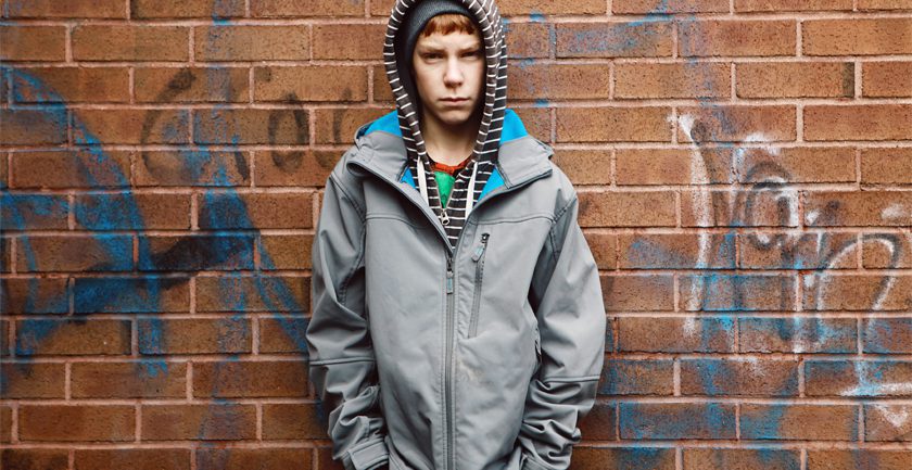 Teenage boy wearing grey jacket and striped hood standing against wall