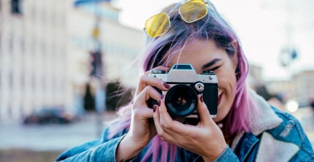Young woman with colourful hair and sunglasses on her head taking a photo with a retro camera.
