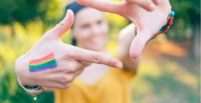 A woman in a yellow top making a hand signal with a rainbow painted on her right hand