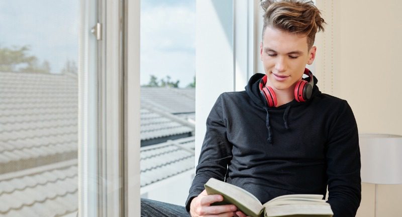 Teenage boy reading a book by a window