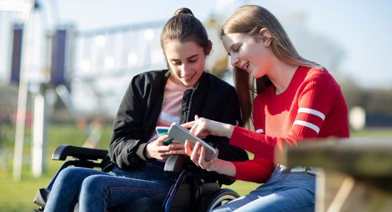 Teenage girl in a wheelchair looking at a mobile phone with a friend