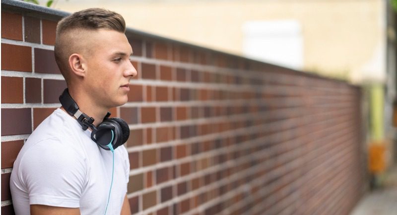 Male teenage with headphones round his neck standing in front of a brick wall