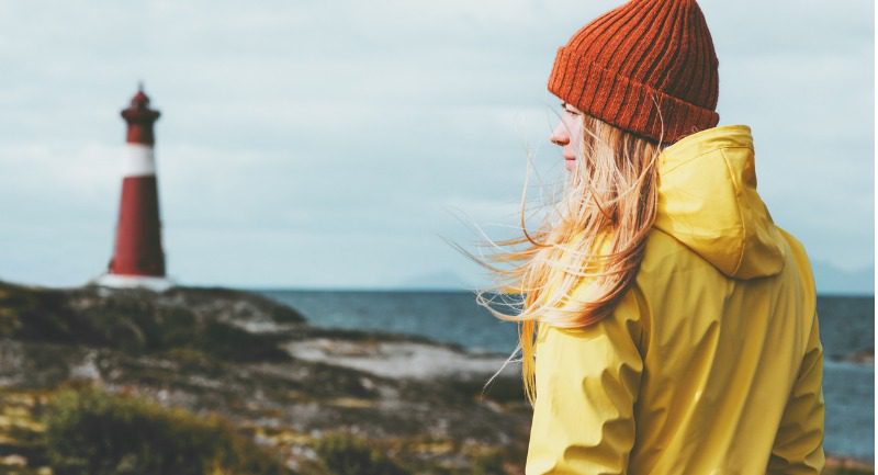 Woman in a yellow jacket and red hat looking at a lighthouse in the distance