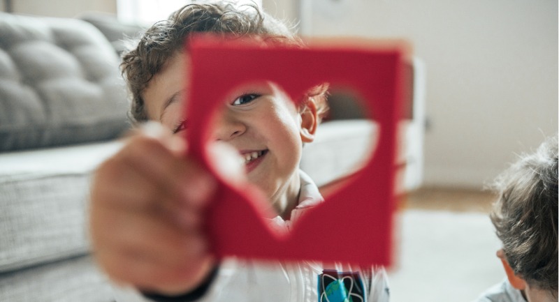 A young child holding up a hollow red heart. Their smile can be seen through the gap.