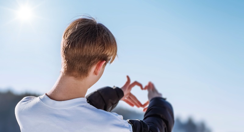 A teenage boy with his back to the camera making a heart gesture with his hands against a bright blue sky