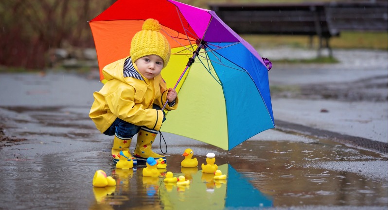 A young boy in a yellow rain jackets holds up a rainbow umbrella. There is a small puddle at his feet with a dozen rubber ducks in it.