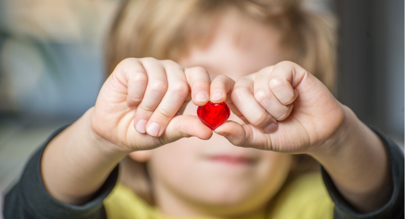 A young girl holds up a small red love heart in her fingers