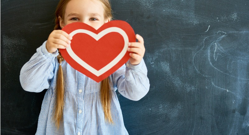 A young girl with long blonde hair holds up a red love heart