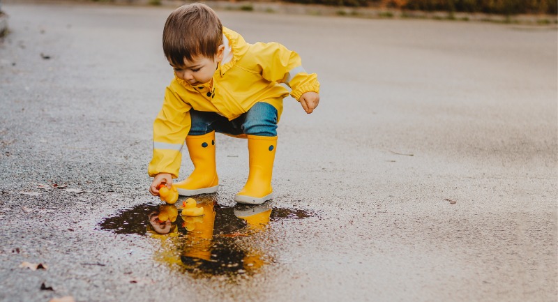 A small child in a yellow rain jacket and yellow wellies plays with a rubber duck in a puddle