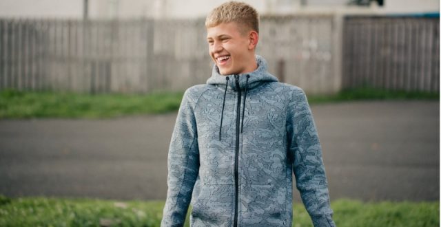 A teenager boy wearing a blue jacket standing smiling in a park