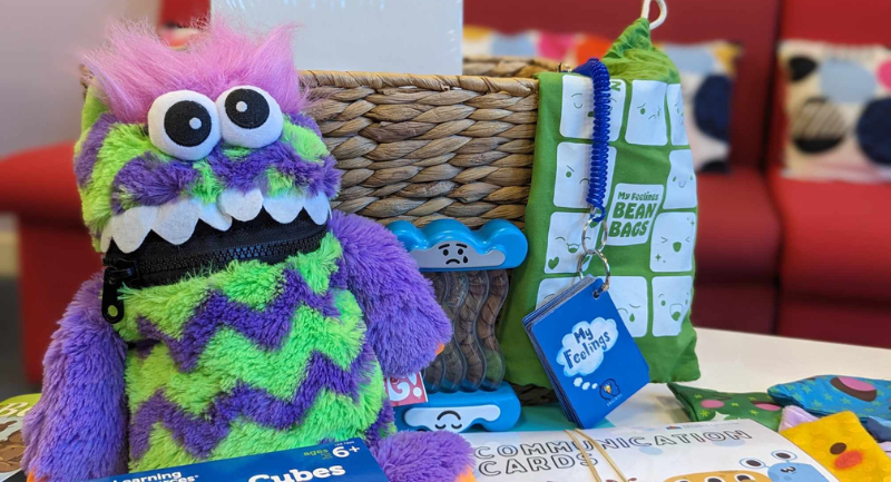 A purple and green monster cuddly toy in front of a straw basket.