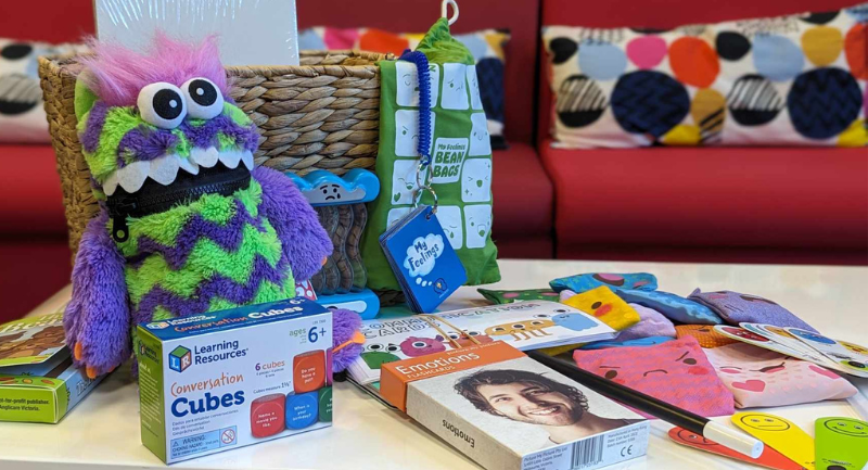 A purple and green monster cuddly toy in front of a straw basket. On the table is a number of other communication resources.