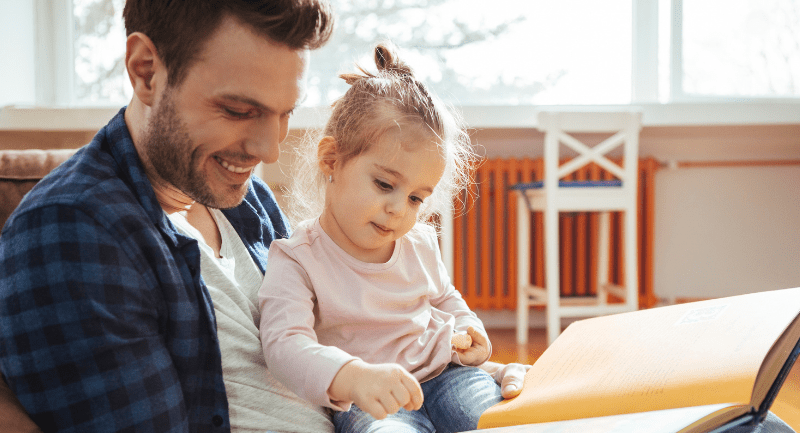 A young daughter sitting on her fathers lap as he reads her a book