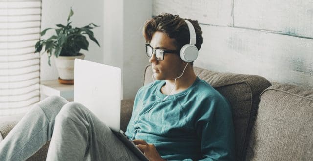 Relaxed young teenager using computer sitting on the sofa at home for play or study. Wearing headphones. One teenager boy listening music in indoor relax leisure activity alone.