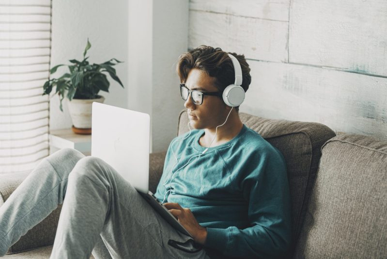 Relaxed young teenager using computer sitting on the sofa at home for play or study. Wearing headphones. One teenager boy listening music in indoor relax leisure activity alone.