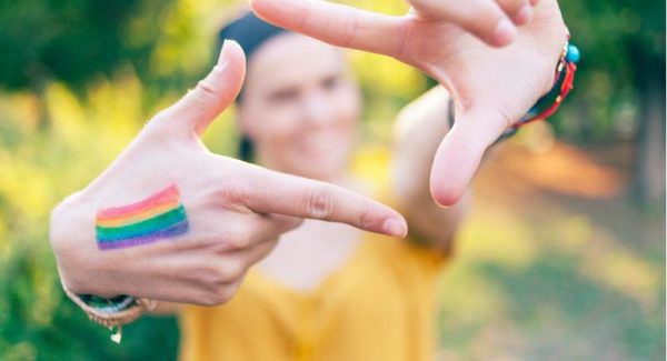 A woman in a yellow top making a hand signal with a rainbow painted on her right hand