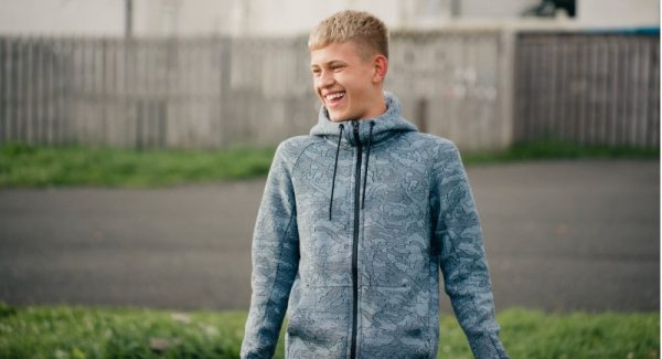 A teenager boy wearing a blue jacket standing smiling in a park