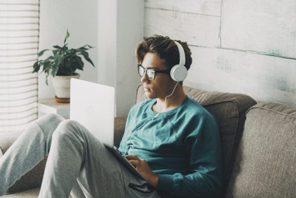 Relaxed young teenager using computer sitting on the sofa at home for play or study. Wearing headphones. One teenager boy listening music in indoor relax leisure activity alone.